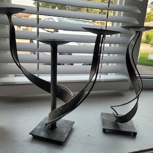 Peut inclure: Deux bougeoirs en métal gris foncé avec une finition texturée. Chaque bougeoir présente une tige centrale verticale fixée sur une base carrée, avec un ruban métallique en spirale qui soutient une plaque circulaire pour poser une bougie.