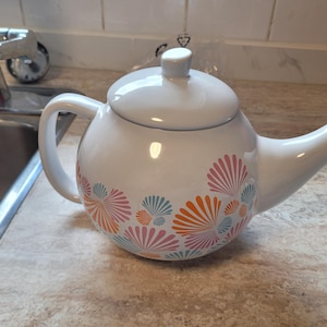 Op de afbeelding: Een witte keramische theepot met een ronde vorm, een bijpassend deksel en een gebogen handvat. De buitenkant is voorzien van een herhalend patroon van gestileerde waaiervormige schelpen in roze, oranje en lichtblauwe tinten rond de basis.