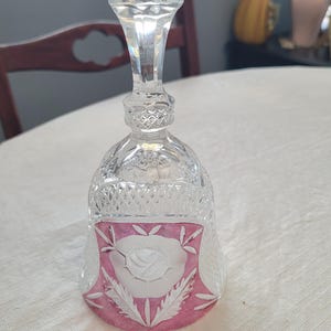 May include: A clear glass bell featuring a faceted handle and a diamond-cut pattern around the upper body. The lower section displays a prominent pink-coloured panel with an etched white rose design surrounded by delicate leaf motifs.