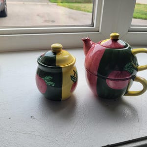 May include: A ceramic tea set consisting of a lidded sugar bowl and a stackable teapot with a cup. The pieces feature a colour-blocked design in dark green, yellow, and pink, decorated with painted red apple motifs and green leaves on the sides.