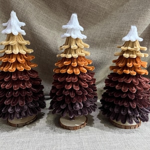 May include: Three velvet Christmas trees in shades of brown, orange, and beige with white felt tops. The trees are on wooden bases.