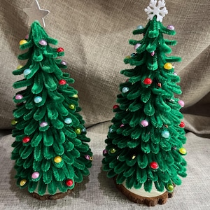 May include: Two green pipe cleaner Christmas trees decorated with colourful beads and topped with a star and snowflake. The trees are on a wooden base.