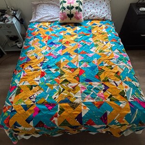 May include: A patchwork quilt featuring a geometric pattern of triangles in vibrant shades of turquoise, mustard yellow, white, and pink. The quilt is topped with a square throw pillow displaying a tufted floral design in pink and green.