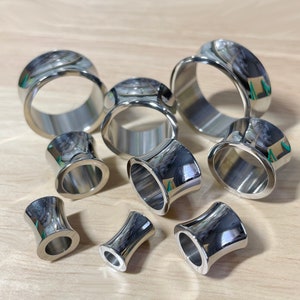 May include: A set of eight silver metal ear gauges of varying sizes. The gauges are all smooth and shiny, and they are arranged in a circle on a wooden surface.