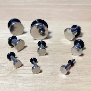 May include: A set of ten silver ear gauges with black o-rings. The gauges are of varying sizes and are arranged on a wooden surface.