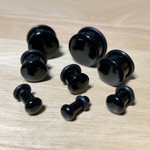 May include: A set of black ear gauges or plugs of different sizes. The gauges are made of a smooth, shiny material and have a rounded shape.