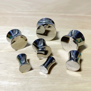 May include: A collection of polished, silver-toned ear plugs in various sizes. The plugs have a flared, cylindrical shape with a flat, circular face. They are arranged on a light-colored wooden surface, reflecting the surrounding light.