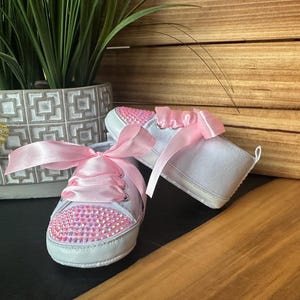 May include: A pair of white baby shoes with pink satin ribbon laces and pink rhinestones. The shoes are on a wooden surface.