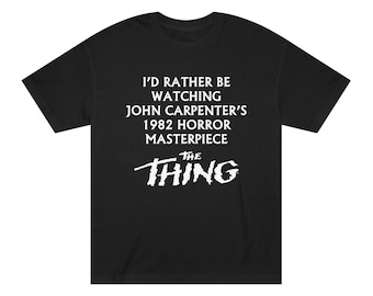 Può includere: Una t-shirt nera a maniche corte con testo bianco che recita I'D RATHER BE WATCHING JOHN CARPENTER'S 1982 HORROR MASTERPIECE seguito dal titolo del film THE THING in un carattere bianco stilizzato e frastagliato sul petto.