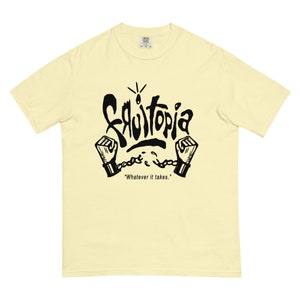 May include: Cream-colored t-shirt with a black graphic design. The design features the word "Fruittopia" in a stylized font, with two fists in shackles and the phrase "Whatever it takes." The shirt has short sleeves and a crew neck.