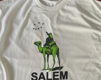 May include: A white crew neck t-shirt featuring a graphic of a green camel with a rider pointing toward a cluster of black stars. Below the camel, the word SALEM is printed in bold black capital letters. The shirt has a standard collar.