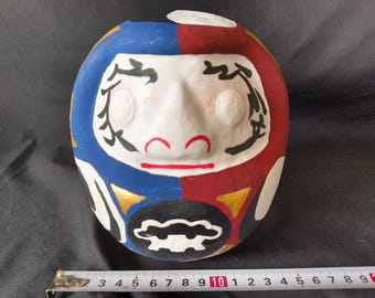 May include: A traditional Japanese lucky charm, known as a "daruma", painted in red, blue, white, and gold. The daruma has a round shape and a painted face with large eyes, a nose, and a mouth. The daruma is a symbol of good luck and perseverance.