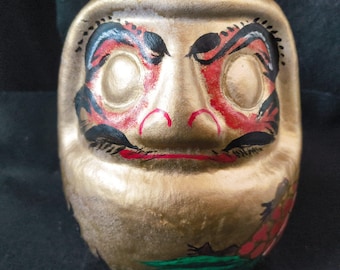 May include: A gold-colored Daruma doll with black, red, and green accents. The doll has a stylized face with large eye sockets, red lips, and black eyebrows. The Daruma doll is a traditional Japanese good luck charm.