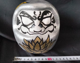 May include: A silver and white Japanese Daruma doll with a black and white painted face and a gold and black painted lotus flower on the bottom.