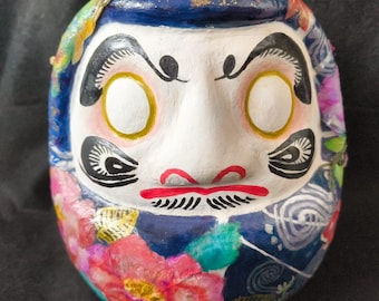 May include: A Daruma doll with a white face, gold-rimmed eye sockets, and a red mouth. The doll's head is painted with a blue background and floral patterns in pink, red, and yellow. The doll has black eyebrows and a mustache.