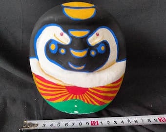 May include: A round, white, black, blue, red, and yellow painted ceramic figurine with a stylized face and a red and yellow striped design around the bottom.