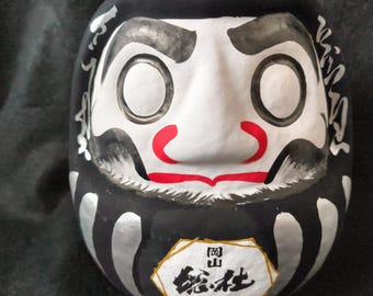 May include: A black Daruma doll with a white face, red lips, and a black beard. The doll has large, open eye sockets and silver accents. The bottom of the doll has a label that reads "SOJA BLACK" in English.