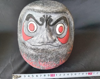 May include: A gray ceramic daruma doll with red and black painted details. The doll has a fierce expression with red eyes and a black mustache. The doll is decorated with red and black flames.
