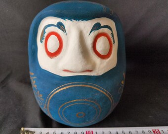 May include: A blue and white Japanese Daruma doll with red eyes and gold accents. The doll is round and has a traditional design.