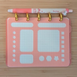 May include: A pink notepad with gold rings and a pink pen. The notepad has a grid pattern and various shapes. The pen is pink and white with the word "Enjoy" printed on it. The notepad and pen are on a wooden surface.