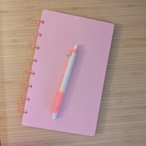 Puede incluir: Un cuaderno rosa con encuadernación en espiral y un bolígrafo rosa y blanco a juego. El bolígrafo tiene un agarre rosa y un cuerpo blanco. El cuaderno es rectangular y tiene una superficie lisa.