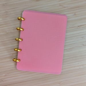 Może przedstawiać: Różowy notatnik ze złotymi pierścieniami do oprawy. Notatnik jest prostokątny z zaokrąglonymi rogami. Tło to jasnobrązowa drewniana powierzchnia.