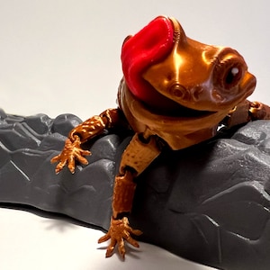 May include: A gold-colored, articulated toy lizard with a red tongue, perched on a gray rock. The lizard has a jointed body and limbs, allowing for various poses.