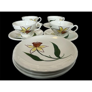 May include: A set of vintage white ceramic dishes with a floral design. The set includes teacups, saucers, and plates. Each piece features a yellow and brown flower with green leaves. The dishes are arranged on a black surface.