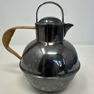 May include: A vintage silver-toned metal pitcher with a woven handle and a black metal handle on top. The pitcher has a rounded body and a spout for pouring. The woven handle is tan in color.