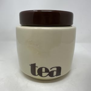 May include: Cream-colored ceramic tea canister with a dark brown lid. The word "tea" is printed in a dark brown font on the front of the canister. The canister has a rounded shape.