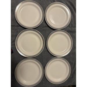 May include: Six white ceramic plates with a black and gray striped border. The plates are arranged in two rows of three on a gray surface. The plates are round and appear to be in good condition.