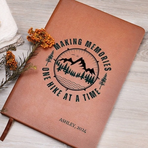 May include: Brown leather journal with a compass design and the text "Making Memories One Hike At A Time". The journal is personalized with the name "Ashley" and the year "2024".