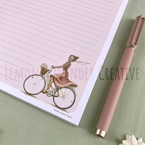 May include: A pink notepad with lined pages and a vintage illustration of a woman riding a bicycle. A matching pink pen rests beside the notepad. The notepad illustration includes the text "Lemon Lavender Creative".
