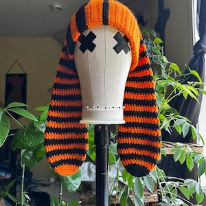 May include: A handmade orange and black striped crochet hat with long ear flaps. The hat is displayed on a mannequin head with black tape covering the eyes. The hat is a unique and stylish accessory.