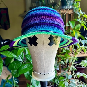 Puede incluir: Un sombrero de ganchillo hecho a mano y colorido con ala ancha. El sombrero presenta rayas horizontales en tonos de azul, morado y verde. El ala está bordeada de verde y amarillo. El sombrero se exhibe en una cabeza de maniquí.