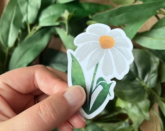 May include: A white daisy sticker with a yellow center and green leaves. The sticker is held by a hand against a backdrop of green foliage. The daisy is a simple, yet elegant floral design.