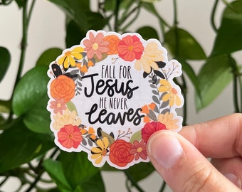 May include: A round sticker with a floral wreath design. The sticker features the text "FALL FOR Jesus HE NEVER leaves" in black script. The wreath includes orange, yellow, and peach-colored flowers and green leaves.