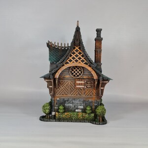 May include: A detailed miniature house with a dark gray roof, wooden walls, and a brick chimney. The house features a decorative arched window, a small stone base, and miniature green bushes. The overall design is reminiscent of a fairytale cottage.