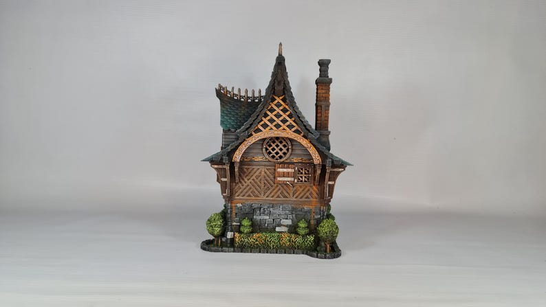 May include: A detailed miniature house with a dark gray roof, wooden walls, and a brick chimney. The house features a decorative arched window, a small stone base, and miniature green bushes. The overall design is reminiscent of a fairytale cottage.
