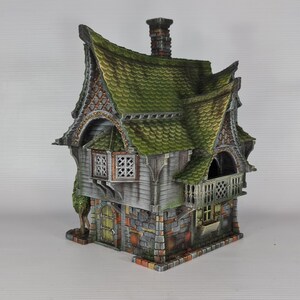 A miniature fantasy house model, with a green roof, gray siding, and stone foundation, features a balcony with a railing and a small green tree on the left side.