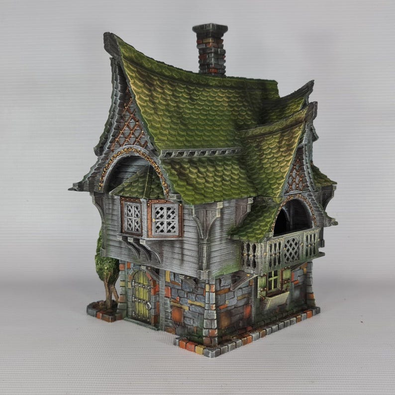 A miniature fantasy house model, with a green roof, gray siding, and stone foundation, features a balcony with a railing and a small green tree on the left side.