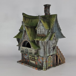 A miniature fantasy house model, with a green roof, gray siding, and stone foundation, features a balcony with a railing and an exterior staircase on the right side.