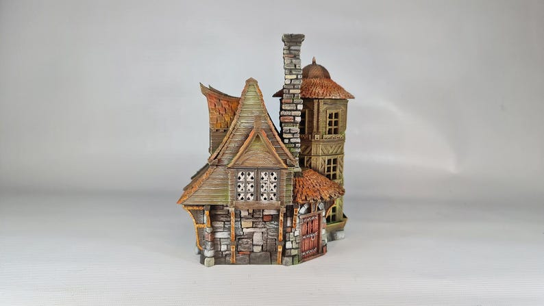A painted miniature of a two-story fantasy building with a red-orange tiled roof, stone foundation, and wooden walls, viewed from the left side, showing a tall chimney.