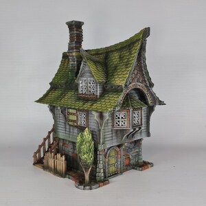 A miniature fantasy house model, with a green roof, gray siding, and stone foundation, is viewed from the front-left, showing a small green tree, a fence, and an exterior staircase.