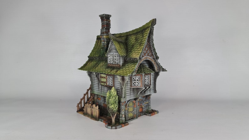 A miniature fantasy house model, with a green roof, gray siding, and stone foundation, is viewed from the front-left, showing a small green tree, a fence, and an exterior staircase.