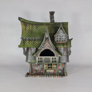 A miniature fantasy house model, with a green roof, gray siding, and stone foundation, is viewed from the front, showing a central balcony and a small window below.