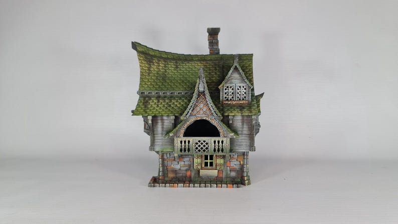 A miniature fantasy house model, with a green roof, gray siding, and stone foundation, is viewed from the front, showing a central balcony and a small window below.