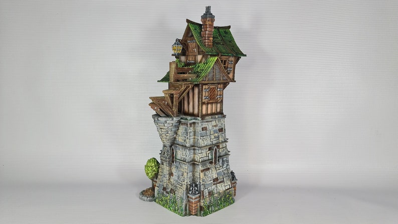 May include: A miniature model of a stone tower with a wooden house built on top. The house has a green roof and a small balcony. The tower is decorated with stonework and has a small tree growing at the base.