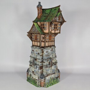 May include: A miniature model of a stone tower with a wooden house perched on top. The house has a green roof and brown wooden walls with windows. The tower is made of gray stone with a brown brick base.
