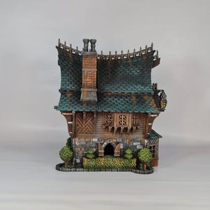 May include: A detailed miniature house with a green tiled roof, wooden siding, and stone accents. The house features a chimney, small windows, and decorative trim. Two small green bushes flank the front of the house.